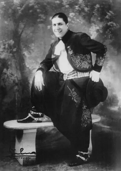 The image appears to be a vintage photograph of a person dressed in traditional mariachi attire. The individual is seated on a stone bench, wearing a charro suit, which includes decorative elements and a wide-brimmed hat. The background features a scenic, slightly blurred setting, suggesting it might be an outdoor scene. The overall style and presentation indicate it may date back several decades.

Image Credits: Wikimedia / Unidentified photographer / Public domain