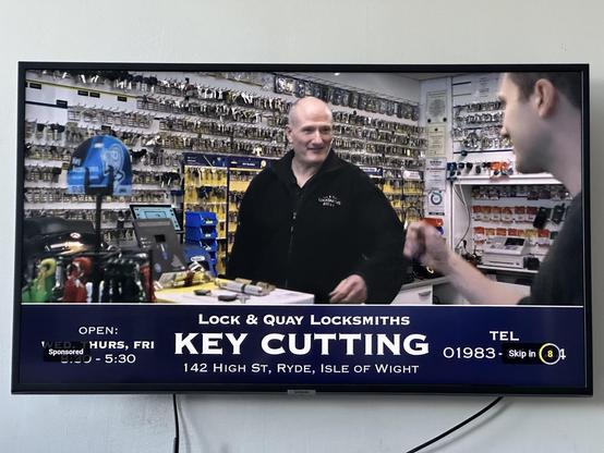 Tv set mounted on a wall with an advert showing lock and quay locksmith key cutting in ryde high street. It has a balding man at the front counter of a key cutting shop with lots of key blanks hanging on the wall behind him
