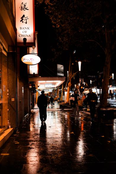 A lone pedestrian walks away, silhouetted against shop lights and the glare of a wet city sidewalk at night
