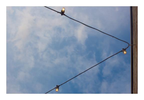 This photograph captures a clear blue sky lightly brushed with wispy white clouds. Two black cables stretch diagonally across the image, forming a triangular shape against the sky. Each cable has small glowing lightbulbs attached at intervals, emitting a soft, warm yellow-orange light. The top right edge of the image includes the corner of a wooden building wall, slightly out of focus, adding a sense of place. The overall mood is calm and minimalist, with strong lines and gentle contrast between the structured cables and the softness of the clouds. The glowing bulbs subtly guide the viewer’s attention across the frame.