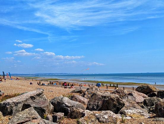 A panoramic view of Worthing beach on a sunny day. In the foreground, large, grey, weathered rocks are scattered across the shingle beach. Beyond the rocks, the beach transitions to smaller pebbles where several individuals are spread out, relaxing and enjoying the sunshine. Towards the horizon, the calm, blue sea stretches out, meeting the lighter blue sky with scattered white clouds. A long, faint landmass is visible in the distance across the water. A colourful kite with an orange tail is flying high in the sky. To the left, a few beach flags or banners are visible, adding splashes of colour to the scene. The overall impression is one of a peaceful, pleasant British seaside day.