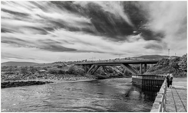 Black and white image depicting a river scene with a bridge overhead. Two people stand near the water's edge on a concrete pier, while rocky terrain and hills are visible in the background under a partly cloudy sky.