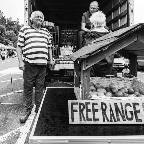Black and white photo of a local market scene in Skibbereen, Co Cork, showing a man in a striped shirt standing beside a display of eggs labelled "FREE RANGE" in front of a van, with two other people engaged in conversation near egg crates in the background.