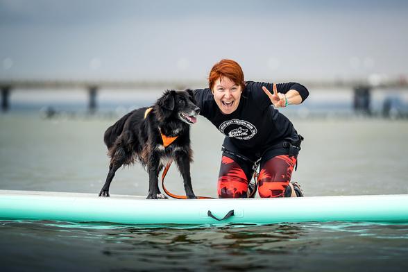 Meine schwarze Border-Collie-Mix-Hündin, Bounty, auf einem SUP-Board auf der Ostsee. Ich knie auf dem Board und mache das Peace-Zeichen Richtung Kamera, schaue lächelnd in die Kamera. Bounty steht klitschnass auf dem Board und schaut mich fragend an.