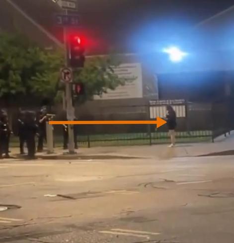 Stop image from video showing a young woman just standing, facing several armed men less than 5 meters/15 f away, one rifle pointing towards woman.
