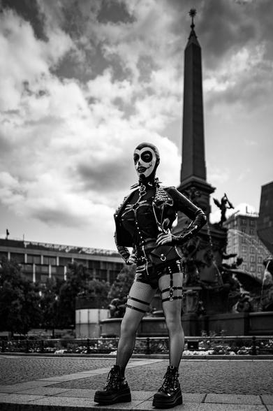 The image captures an adult in a latex suit and mask standing confidently with their hands on their hips. In the background, a monument and a fountain add to the urban landscape. The foreground features a patterned ground, providing a stark contrast to the sky.