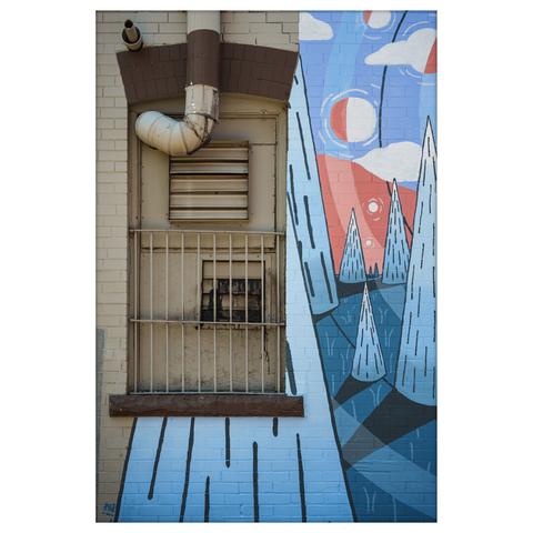 A photograph of a building exterior featuring a heavily weathered and barred window with a rusted exhaust fan behind metal bars. Above the window, an old ventilation pipe curves outward and up, attached to a beige wall made of painted bricks. The right side of the wall is covered in a vibrant mural: a stylized landscape with triangular trees, flowing lines, floating clouds, and large red and white circular shapes resembling suns or moons. The mural's palette includes sky blue, navy, coral red, and white, contrasting sharply with the dull, industrial elements of the window and pipe. The image captures the tension between decay and creativity in an urban setting.
