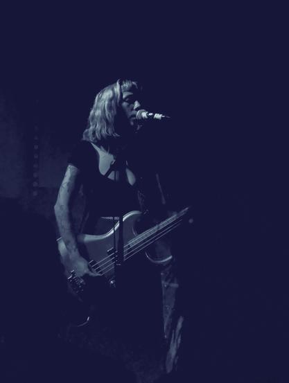 A young blonde woman is playing a bass guitar in front of a microphone. She is wearing a black top with short sleeves, exposing her right arm where she shows some tattoos. The scene is very dark and the musician is only partially visible from her right side, as if she were devoured by the darkness. The picture is black and white but with a slight blue tone.