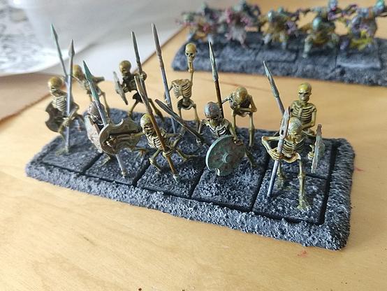 A unit of ten skeleton miniatures on a movement tray. The skellies are armed with tarnished bronze spears and shields. One in the back is curious and holds up his head to see over the row before him.