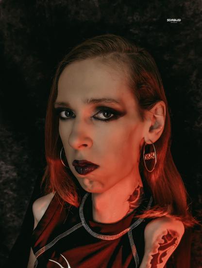 Porträt von mir im empowernden, radikalen Look: rote Haare, dunkles, ausdrucksstarkes Augen-Make-up, dunkler Lippenstift. Große silberne Creolen mit dem Schriftzug "Disobey", schwarze Kleidung mit silbernen Nähten, markante Tattoos an Hals und Schulter. Das rote Licht und der dunkle Hintergrund setzen ein klares Statement: Sichtbarkeit. Unangepasst. Laut. Queer, behindert, schön — trotz und wegen eines Systems, das genau diese Bilder selten zeigt.