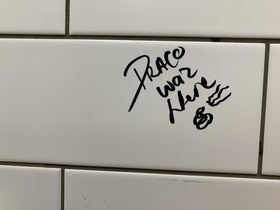 Black marker graffiti on white wasroom tile stating "Draco waz [sic] Here" followed by a scribble and three wavy lines, perhaps indicative of a New Wave haircut or a poorly-drawn rendering of an emoji.