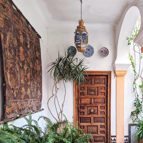 An enclosed courtyard or patio with a distinctly Moorish aesthetic. A rich brown wooden door with intricate geometric paneling and a brass knocker is centrally located. Above the door, a detailed blue and gold metal lantern hangs from the ceiling. To the left of the door, a large, ornate tapestry with a floral pattern in browns and oranges is hung on the white wall. Below the tapestry, a vibrant green fern spills out of a planter, and a taller plant with spiky leaves stands in a pot beside the door. To the right of the door, three decorative blue and white ceramic plates are mounted on the wall. An arched doorway with a warm orange-yellow frame and climbing vines leads to another area on the right. The ground is a mix of white and blue patterned tiles near the door.