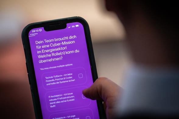 Auf einem Smartphonebildschirm wird eine Frage an einen Spielteilnehmer gerichtet: "Dein Team braucht dich für eine Cybermission im Energiesektor! Welche Rolle(n) kannst du übernehmen?"