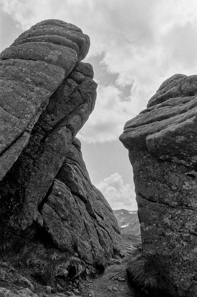 This black-and-white photograph, taken in the Carpathian Mountains, captures a dramatic natural rock formation. Two massive rock walls rise on either side of a narrow path, creating a striking visual corridor that leads the eye toward the distant mountains visible through the gap. The rocks display weathered textures and layered structures, with tufts of grass growing at their base. Above, a partly cloudy sky adds depth and atmosphere to the scene. The image has a timeless, almost monumental quality, emphasizing the raw beauty and ruggedness of the Carpathian landscape