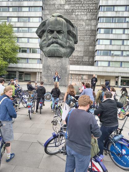 Ein Mensch mit Mikrophon steht auf dem Sockel vor dem Karl-Marx-Monument und spricht zu einer Gruppe von ca. 20 Radfahrern.