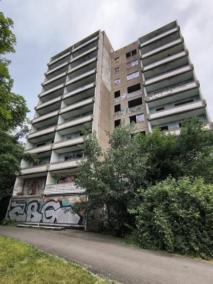 Leerstehender und mit Graffiti besprühter 10-geschossiger Plattenbau mit leeren Fenstern und vielen Balkons, davor Büsche und ein asphaltierter Weg.