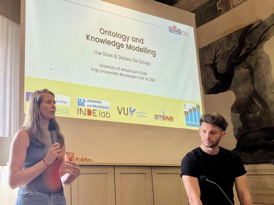 Lise Stork (left) and Stefano de Grigoris (right) standing in front of the projection showing their cover slide for the Ontology and Knowledge MOodelling tutorial