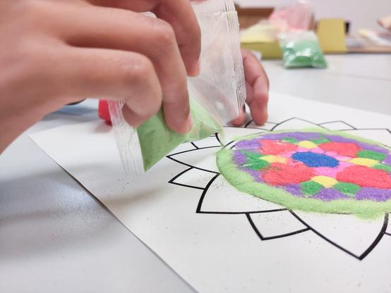 A close-up image of hands creating a colorful sand art design. The hands are pouring green sand from a small plastic bag onto a mandala pattern drawn on a white sheet of paper. The mandala design is partially filled with various colors of sand, including red, blue, purple, yellow, and green. In the background, there are other bags of colored sand and art supplies on a table.
