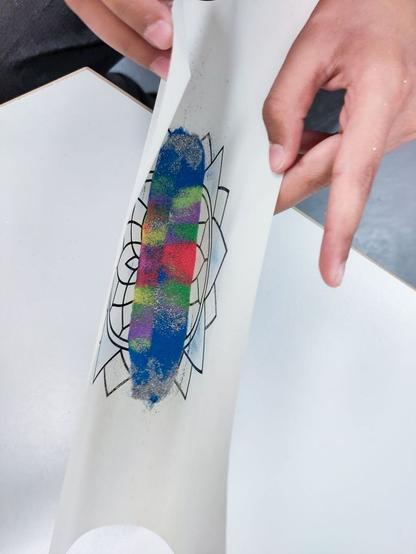 A close-up of hands holding a sheet of paper with a partially completed sand art design. The design features a geometric outline, and colored sand in blue, green, pink, and red has been applied to the center area.
