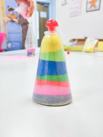 A finished sand art bottle sits on a table. The cone-shaped bottle is filled with neatly layered colored sand in yellow, green, blue, pink, red, and glitter, and is sealed with a red cap. In the background, another person is seen working on a similar sand art project.