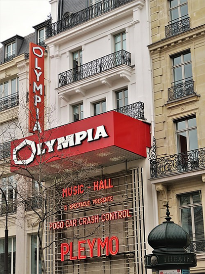 Das Foto zeigt die Fassade des Pariser Konzertsaals „Olympia“. Es handelt sich um ein mehrstöckiges Gebäude mit klassischer Fassade und schwarzen Eisengeländern vor den Fenstern. Ein großer roter Schriftzug mit weißem Logo prangt über dem Eingang: OLYMPIA. Seitlich davon ein senkrechter Leuchtschriftzug mit dem Wort OLYMPIA in weißen Großbuchstaben auf rotem Hintergrund.