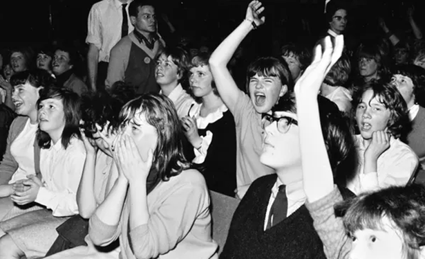 Ein Schwarz-Weiß-Foto aus den 1960er-Jahre zeigt eine Gruppe junger Menschen in einem Konzertsaal. Die meisten im Publikum sind Mädchen im Teenageralter. Viele wirken aufgeregt und emotional: Einige reißen die Arme hoch, andere schreien, klatschen oder halten sich die Hände vor den Mund. Eine Person trägt eine große Brille und schaut konzentriert nach vorn. Die Jugendlichen sitzen eng beieinander in mehreren Reihen, im Hintergrund stehen einige Erwachsene. Die Szene vermittelt einen Moment intensiver Begeisterung und kollektiver Euphorie.