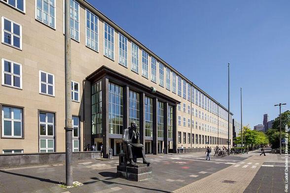 Hauptgebäude der Universität zu Köln mit breiter Fensterfront und Eingangstüren. Vor dem Gebäude steht eine Statue eines sitzenden Mannes (Albertus Magnus). Einige Passanten gehen vorbei, Fahrräder sind abgestellt. Bei klarem Himmel.