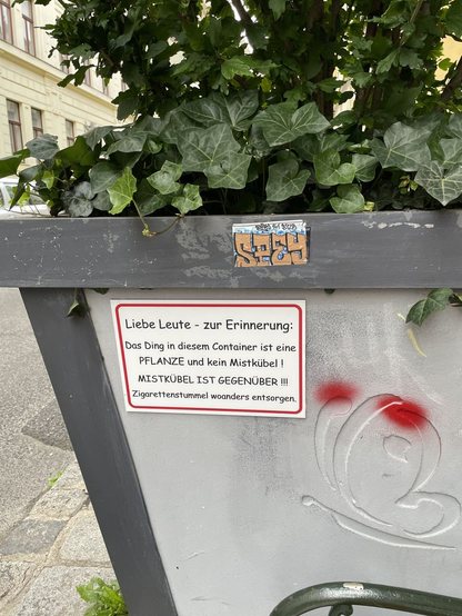 Ein Schild hängt am Riesenblumentopf in der Stadt wo draufsteht: "Liebe Leute - zur Erinnerung: Das Ding in diesem Container ist eine PFLANZE und kein Mistkübel! MISTKÜBEL IST GEGENÜBER!!! Zigarettenstummel woanders entsorgen