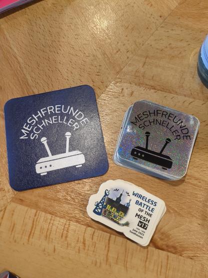 Photo of two packs of stickers and a drink coaster on a wooden surface.
The coaster and one sticker pack have the logo of "Meshfreunde schneller" (a WiFi router with two antennas and the text "meshfreunde schneller" above.
The second sticker pack has the logo of the battlemesh v17 (a antenna tower and an old house)