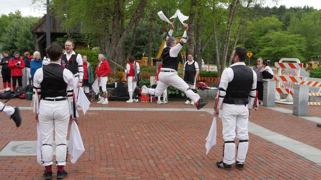 Four morris dancers (plus the foot of a fifth) with white shirts and trousers and black vests, decorated with multicolor ribbons, dancing on a brick paved surface. Three are standing and one is leaping high with arms raised. Behind them an audience including some other morris dancers.