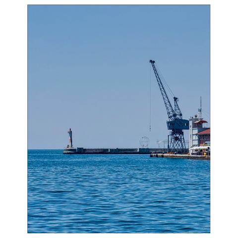 The image shows a serene coastal scene with a calm sea under a clear blue sky. On the left, a small lighthouse stands at the end of a pier. To the right, a large industrial crane is visible, likely part of a port facility. In the background, there is a spherical metal structure, adding to the industrial feel of the scene.