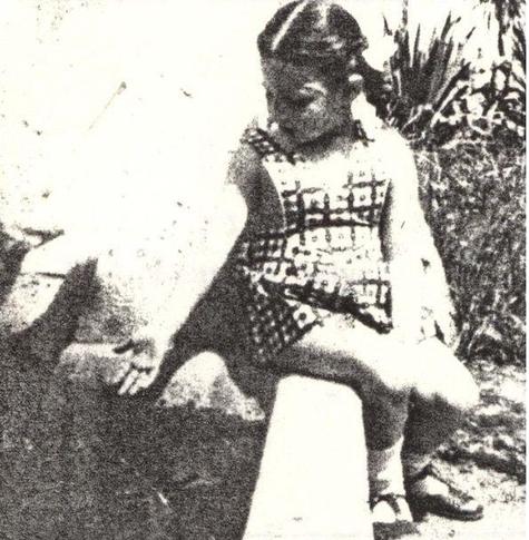 A young girl in a patterned dress is sitting outdoors on a sunlit day, focusing intently on something in their hands, surrounded by a natural, grassy environment.