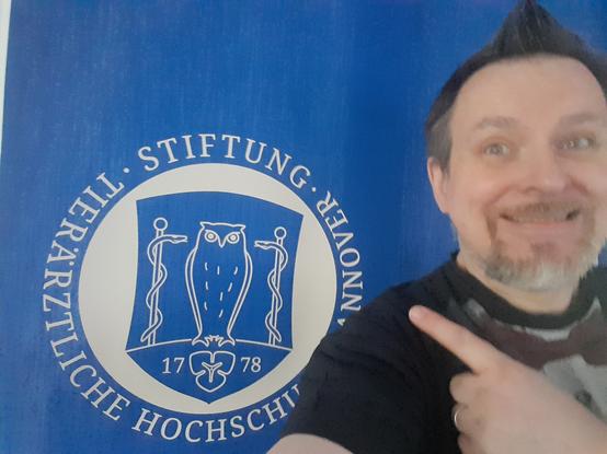 Foto, selfie, ich grinse vor blauem hintergrund und zeige auf das Logo auf dem blauen hintergrund der tierärztlichen hochschule hannover