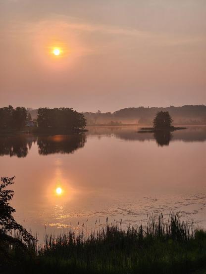 The image depicts a serene lakeside scene at sunrise. The sun is low in the sky, casting a warm, golden glow across the scene. Its reflection is visible on the calm surface of the lake, creating a mirror-like effect. The sky is painted with soft hues of pink and orange, blending into a light blue near the horizon. In the foreground, there is a strip of greenery, including tall grasses and a few trees, framing the view. The lake is dotted with small islands, one of which is more prominent and has a cluster of trees. The misty atmosphere adds a sense of tranquility to the scene. The overall composition is balanced, with the sun, islands, and reflections creating a harmonious visual.