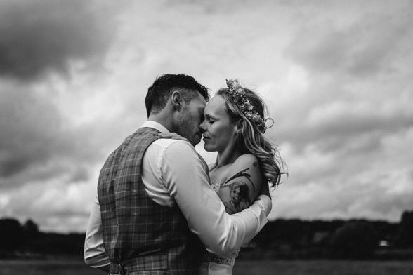The image is a black and white photograph. It captures a tender moment between a bride and groom standing close together, their faces touching in a loving embrace. The groom is dressed in a vest and a white shirt, while the bride is adorned in a dress and a floral crown. The background features a cloudy sky and a field, suggesting an outdoor setting. The black and white nature of the photo adds a timeless quality to the scene. The couple's posture and closeness convey a sense of intimate connection and joy on their special day.