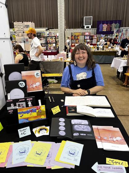 Artist portrait on comics fair with comics and zines on a black table, in the background people buzzing about the fair, stands with artworks, stickers and gadgets