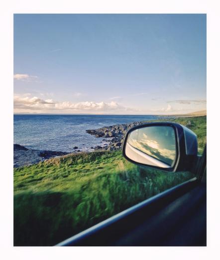 The photo is of the view from inside a moving car, looking out the side window and the side mirror. The view is of a coastaline. There a stretch of dark-green grassy land next to a rocky shoreline and a calm blue sea that is under a mostly clear sky with a few fluffy clouds near the horizon.