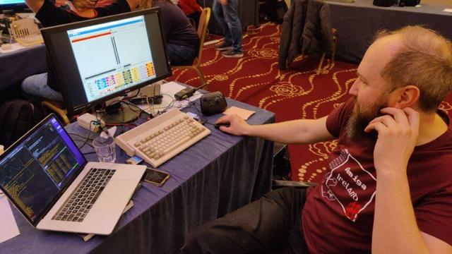 A man with a red shirt. on a chair beside a desk. On the desk there is a laptop and an very old computer with a screen.