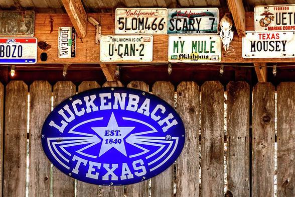 A wall made of wood fence with an oval sign of Luckenbach Texas est 1849 is bellow the header of a rooftop.  On the header are many car tags such as BOZO, U-CAN-2, My Mule, Mousey, and more specialized ones.  Photography by Debra Martz