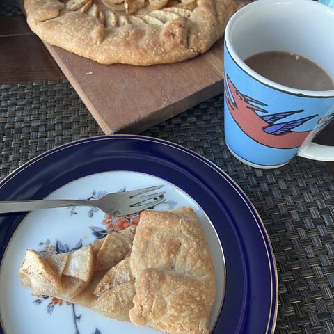 A slice of apple pastry, next to a fork, on a decorated plate. There is a wooden board with more galette, and colorful coffee mug decorated with a cartoon winged dog