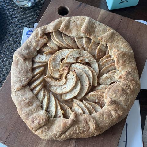 A round apple galette with spiral-arranged apple slices in a toasted crust folded over at the edges, on a wooden cutting board.