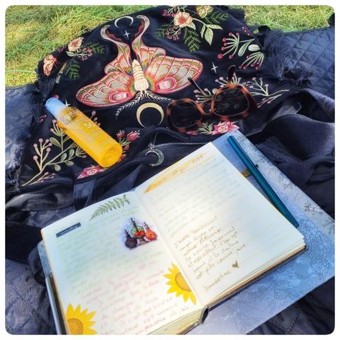 un sac brodé d'un papillon est posé à plat sur une couverture sur l'herbe, une huile solaire, des lunettes de soleil et un beau carnet d'art journal sont élégamment posés autour.