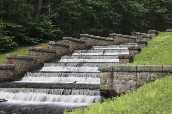 A series of stone steps with flowing water cascading down, surrounded by greenery. The scene features a tranquil waterfall effect with moss and ferns visible on the sides.