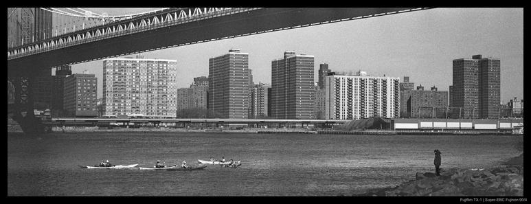 A person stands on a rocky shore with the Manhattan skyline visible across the East River.  A group of four kayaks paddles in the river between them, beneath a large bridge.