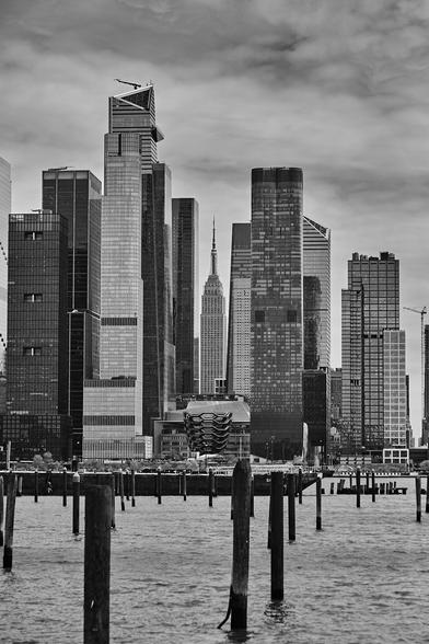 Black and white photo of Manhattan’s west side skyline, with the Empire State Building framed between modern skyscrapers. Wooden pilings rise from the Hudson River in the foreground under a cloudy sky