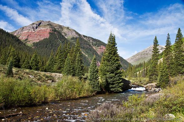 Photo of a shallow creek flowing through a verdant narrow valley with large mountains in the background, on the left with bare, red-layered cliff face; conifers; blue sky with high thin clouds. A bit of a cascade in the creek.
©BosqueBill.com