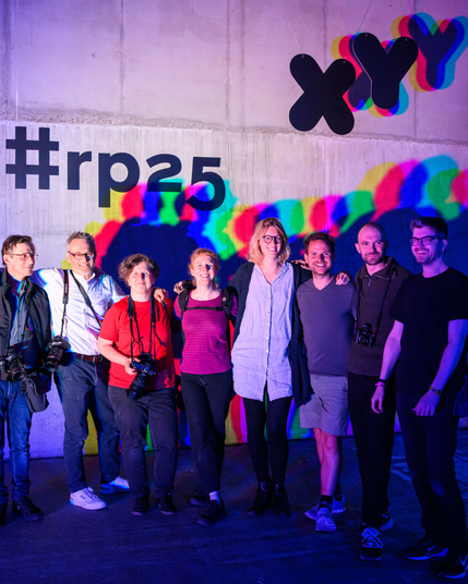 Foto aller Fotograf*innen der republica vor einer Lichtinstallation. Viele von ihnen tragen ihre Kamera bei sich und alle läcjeln in die Kamera. Im Hintergrund ist der Hashtag rp25 zu sehen und die Buchstaben XYZ hängen von der Decke.