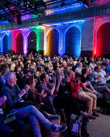 Das Publikum an Bühne zwei klatscht. Menschen sitzen in mehreren Reihen hintereinander. Im Hintergrund sind bunte Lichtstrahlen zu sehen, die die Backsteinwand anstrahlen.
