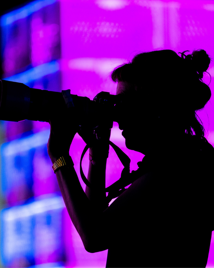 Silhouette von Anne Barth, Fotografin, auf der republica. Sie schaut gerade durch die Kamera und trägt ihre Haare zu einen Dutt. Dahinter sind die bunt angestrahlten Bäckerkisten der republica 2025 zu sehen.