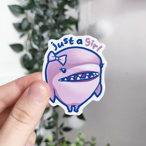 Hand holding up a sticker of a pink shark with lipstick and a ribbon bow titled "just a girl"